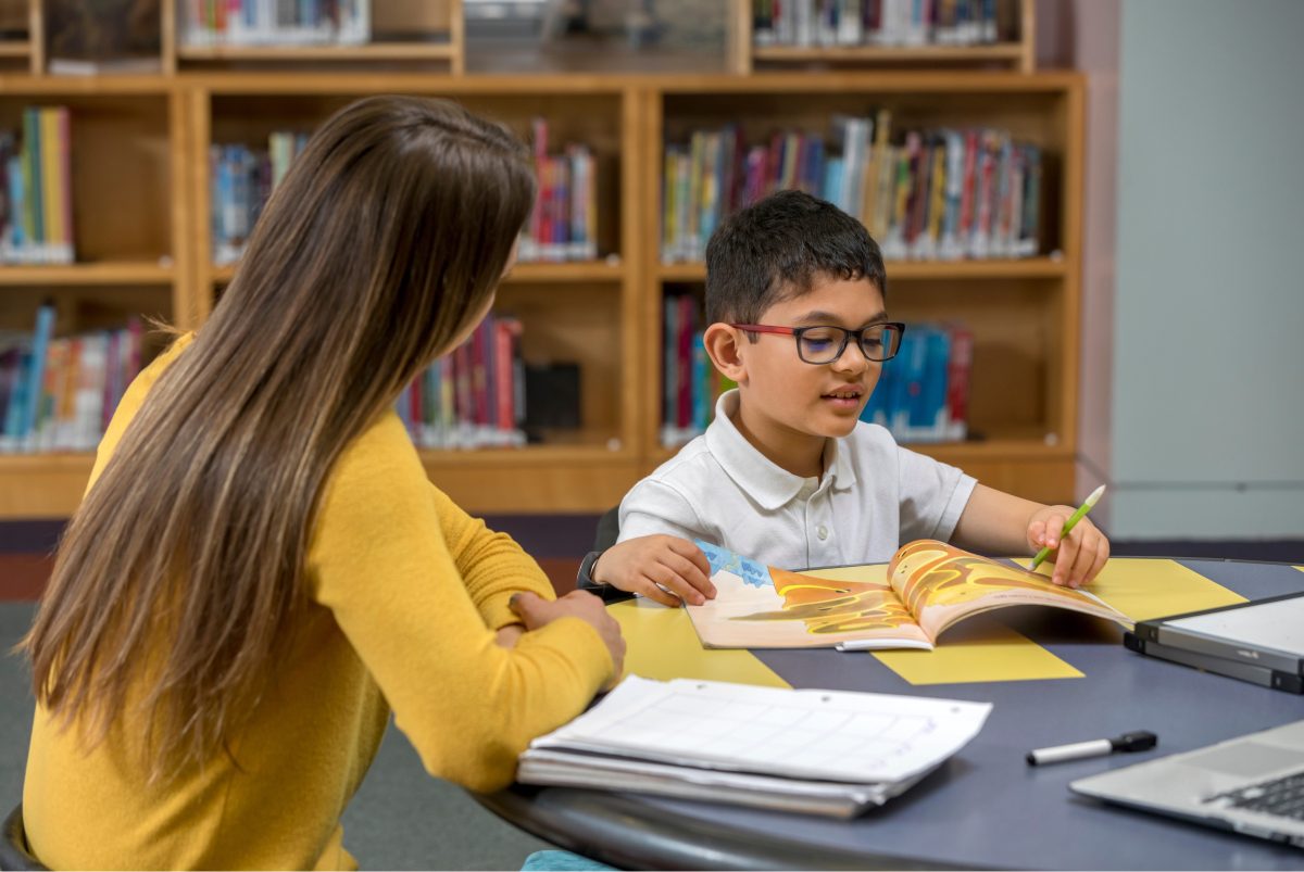 A tutor helps a young boy with glasses read in a school library
