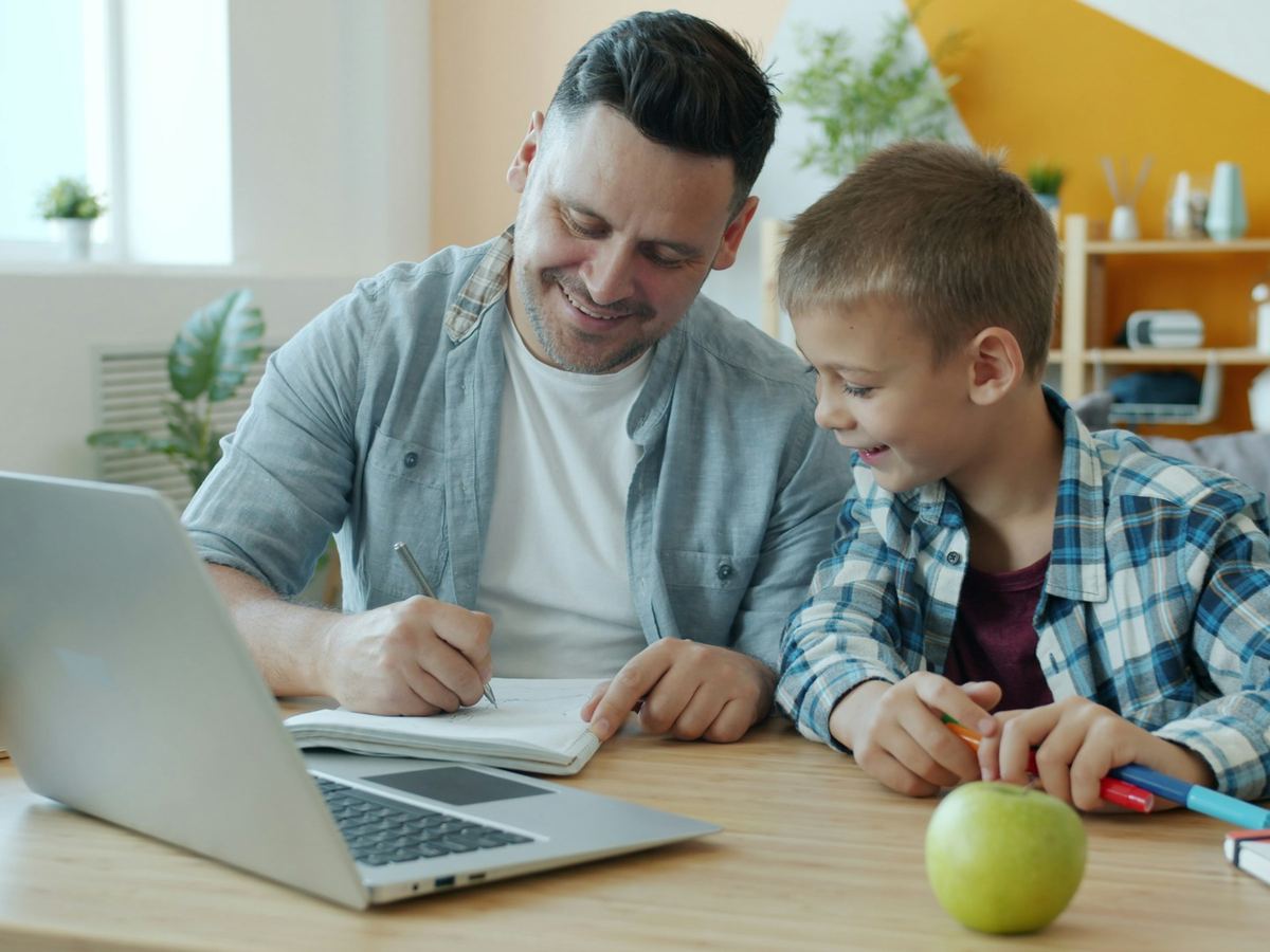A smiling man helps a boy with schoolwork at a table