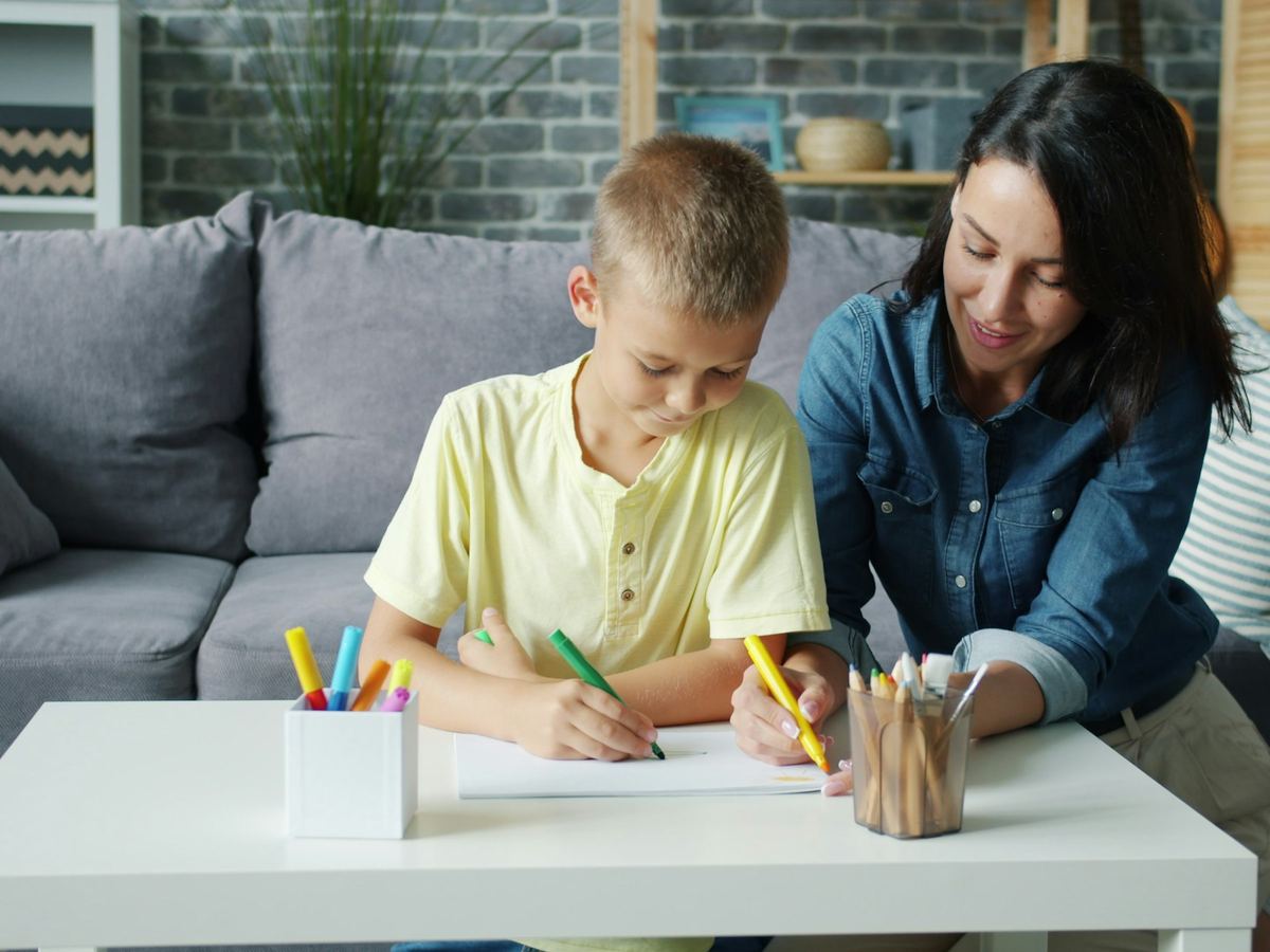 An adult sits beside a boy as he works at a table